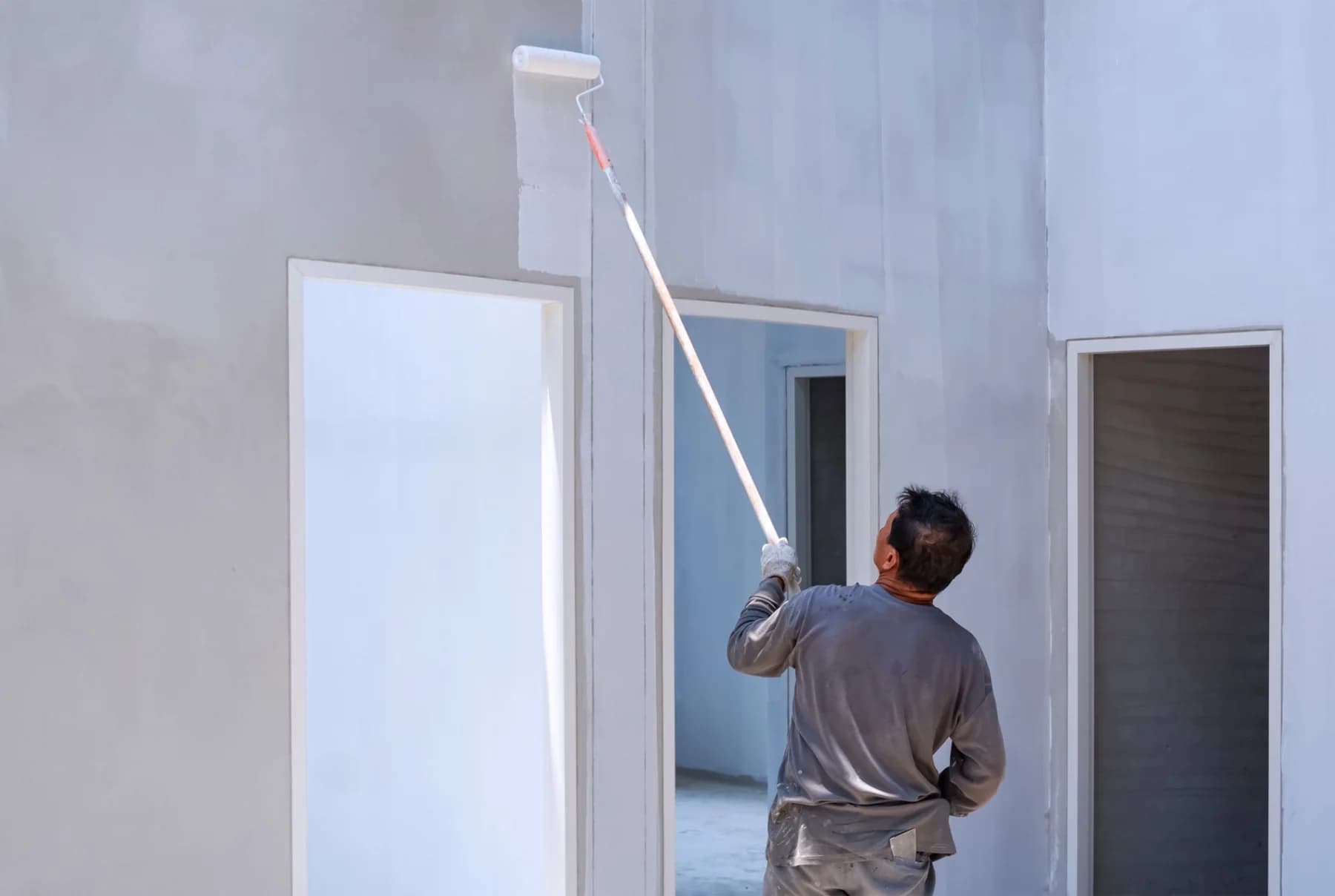 A painter rolling a coat onto a grey wall with a long-handled roller